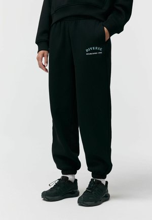 Osoba ubrana w czarne dresy z napisem „DIVERSE Established 1993”, w połączeniu z czarnymi trampkami i białymi skarpetkami, ręce swobodnie opuszczone wzdłuż ciała.