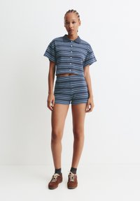 Femme se tenant devant un mur blanc, portant une chemise à manches courtes boutonnée à rayures bleues et un short assorti, avec des chaussures marron.