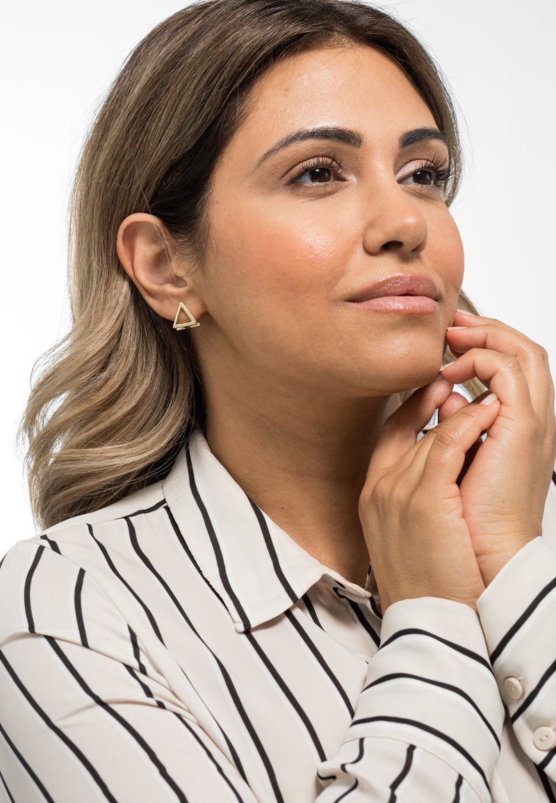 Orecchino a forma di triangolo in tonalità dorata indossato da un modello. L'orecchino presenta un design minimalista con una finitura lucida, abbinato a una blusa a righe.
