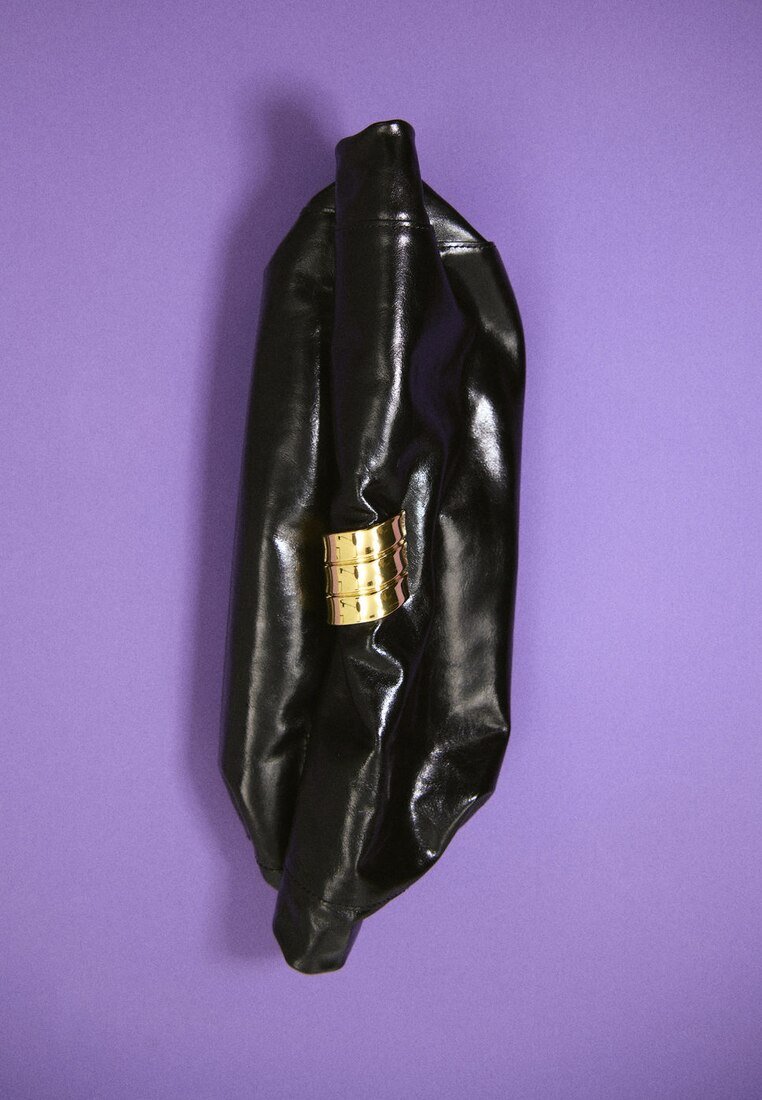 Sac à main en cuir noir brillant avec une large agrafe décorative dorée sur un fond violet uni.