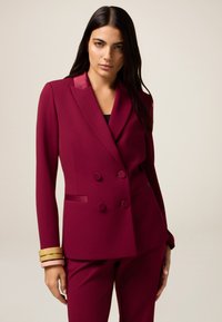 Blazer bordeaux doppiopetto con revers in satin, due tasche frontali e dettagli con bottoni. Abbinato a pantaloni aderenti coordinati. Tessuto morbido.