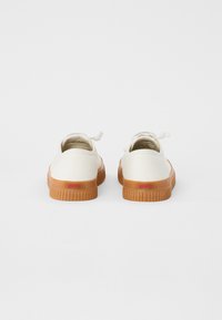 White canvas low-top shoes with a gum rubber sole, featuring red branding on the heel and minimalistic design with a round toe.