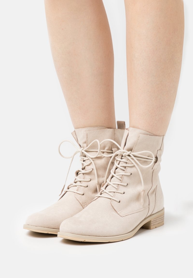 Botas de gamuza beige hasta el tobillo con cordones, usadas sobre piernas desnudas contra un fondo blanco.