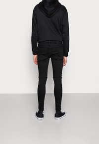 Sudadera negra con capucha y jeans skinny negros vistos desde la parte de atrás, combinados con zapatillas altas negras. Tela suave y diseño ajustado.