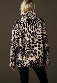 Kobieta ubrana w czarno-beżową kurtkę z polaru w zwierzęcy wzór z wysokim kołnierzem, stojąca na tle jednolitego ciemnobrązowego tła.