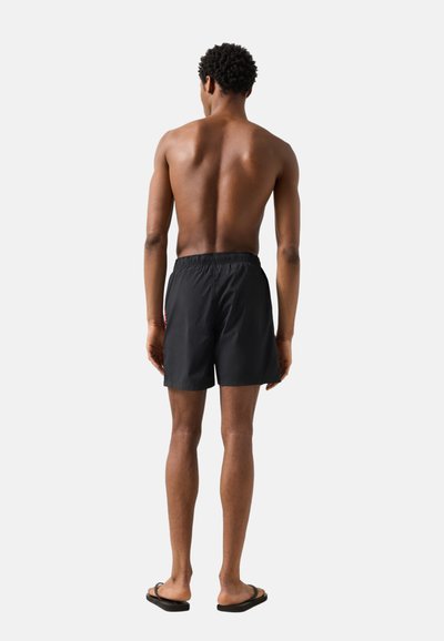 Homme debout, de dos, portant un short de bain noir et des tongs noires, sur un fond uni gris clair.