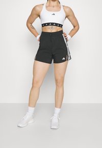 Bílé sportovní podprsenka s černým lemem a logem, zkombinovaná s černými šortkami s bočními pruhy a logem. Vzhled doplňují sportovní ponožky a boty.