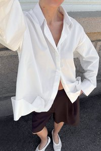 White cotton button-up shirt with a loose fit, folded sleeves, and curved hem. Paired with brown shorts and white woven slip-on shoes.