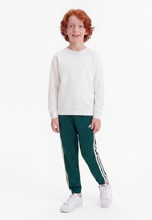 Jeune enfant roux souriant, portant un t-shirt blanc à manches longues, un pantalon de survêtement vert foncé avec des bandes blanches et des baskets blanches.