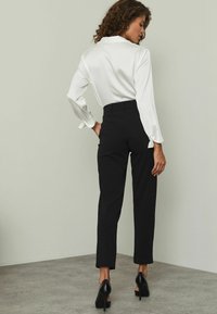 Bílá saténová blouse s dlouhými rukávy, černé propracované kalhoty a černé vysoké podpatky. Hladká textura a čisté linie. Pohled zezadu na jednolitém pozadí.