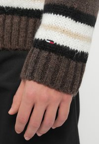 Καφέ πλεκτός μανίκι από πουλόβερ με μαύρες, λευκές και μπεζ ρίγες. Διαθέτει ραβδωτή υφή και μικρή ετικέτα με λογότυπο σε κόκκινο, λευκό και μπλέ.