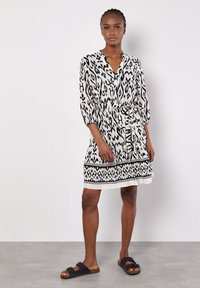 Schwarz-weißer gemusterter Dress mit V-Ausschnitt, drei Viertelärmeln, einem Taillengürtel und dekorativem Saum. Getragen mit dunklen Sandalen.