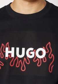 Sweat-shirt noir à col rond avec texte blanc "HUGO" et motifs de flammes rouges sous le texte sur la poitrine.