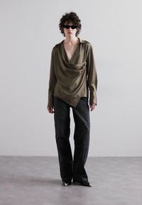 Olivgrünes, locker fallendes Hemd mit tiefem V-Ausschnitt, kombiniert mit lockeren, dunklen Jeans. Das Model trägt schwarze Sonnenbrille und spitze schwarze Schuhe.