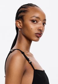 Top negro sin mangas con tirantes ajustables y herrajes plateados, de textura suave. La modelo tiene el pelo trenzado y piercings faciales.