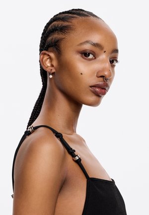 Top negro sin mangas con tirantes ajustables y herrajes plateados, de textura suave. La modelo tiene el pelo trenzado y piercings faciales.