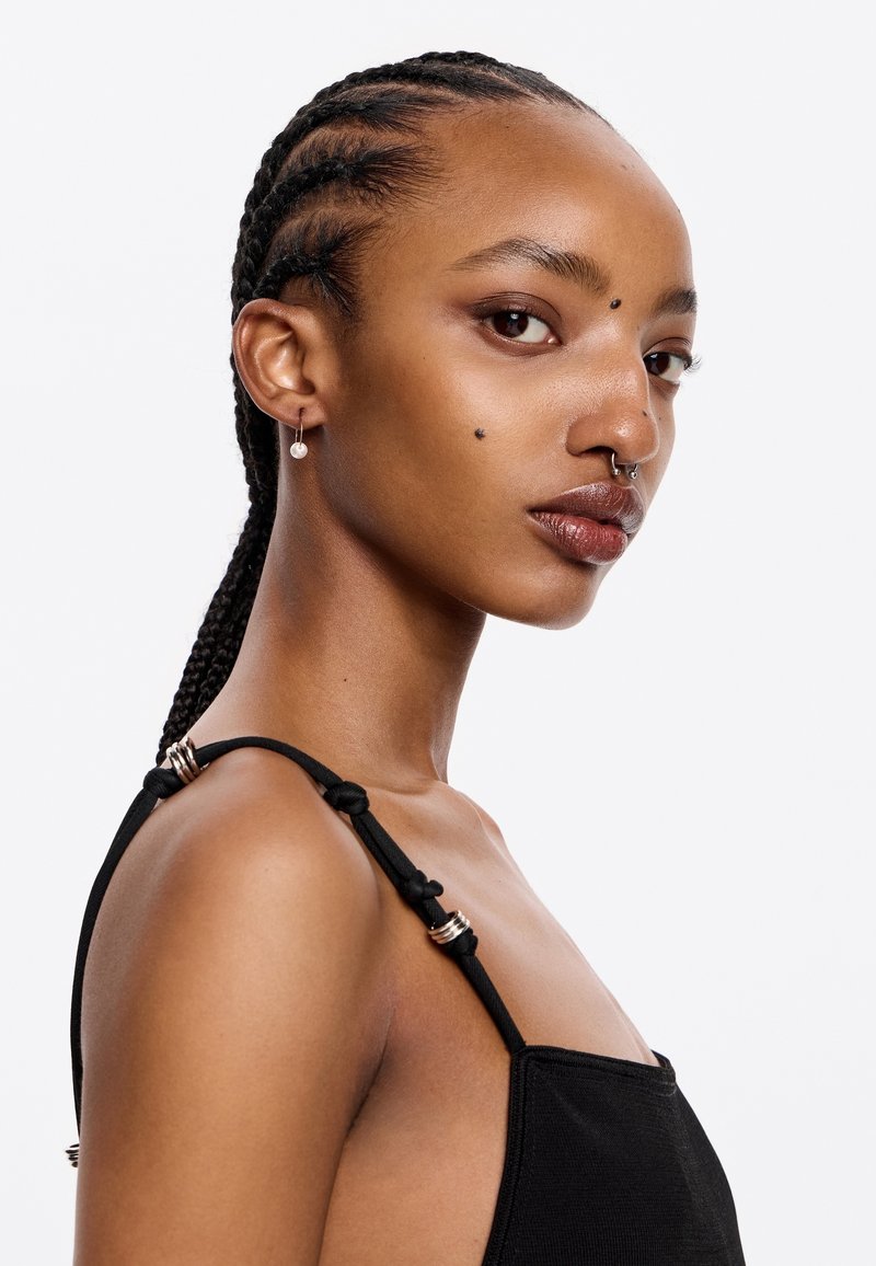 Top negro sin mangas con tirantes ajustables y herrajes plateados, de textura suave. La modelo tiene el pelo trenzado y piercings faciales.