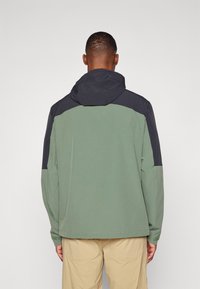 Veste à capuche avec un corps vert et des épaules bleu marine. Présente une coupe décontractée, des manches longues et une texture lisse. Aucuns logos visibles.