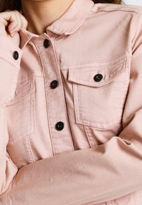 Ljust rosa denimjacka med svarta tryckknappar, två bröstfickor och krage. Tyget har en mjuk, slät textur.