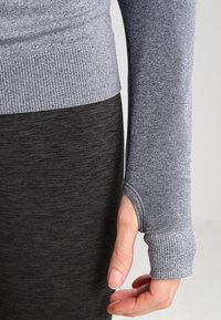 Gros plan sur une main émergeant d'une manche côtelée grise avec un trou pour le pouce, reposant contre une jupe ou un pantalon texturé gris foncé.