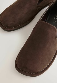 Paire de chaussures en daim marron à enfiler avec des bouts carrés arrondis et des coutures visibles le long des bords sur une surface claire.