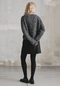 Czarny dziergany sweter z cętkowanym wzorem, zestawiony z czarną spódnicą i czarnymi rajstopami. Modelka stoi z rękami skrzyżowanymi za plecami.