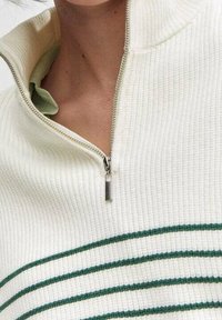 Biały sweter dziany z kołnierzem na połowę zamka, częściowo rozpięty, odsłaniający zieloną koszulę pod spodem z poziomymi zielonymi paskami.