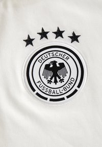 Tecido branco com um emblema redondo de camiseta de futebol preto e branco, apresentando uma águia, o texto "DEUTSCHER FUSSBALL-BUND" e quatro estrelas pretas acima.