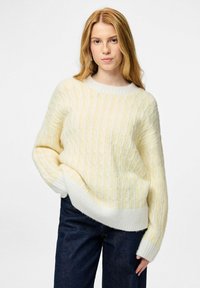 Šviesiai geltonas ir baltas megztinis su pynės raštu, apvaliu kaklu ir ribotu apačioje, derinamas su tamsiai mėlynomis džinsinėmis kelnėmis.