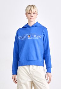 Hanorac albastru dintr-un material din bumbac. Dispune de o glugă cu șnur, buzunar frontal și text grafic alb cu logo. Creat pentru îmbrăcămintea casual.
