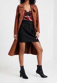 Abrigo largo de gamuza marrón sobre una blusa de encaje de cuadros rojos, combinado con una falda negra con cinturón y botas de tobillo negras con tacones y detalles plateados.