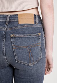 Mörkblå denimjeans med ljusorangea sömmar, standardfickor och en läderetikett på den bakre midjan. Närbild av bakifrån.