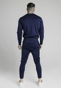 Man med nacktattoo som bär en marinblå långärmad topp och matchande byxor, stående med ryggen mot en enkel grå bakgrund.