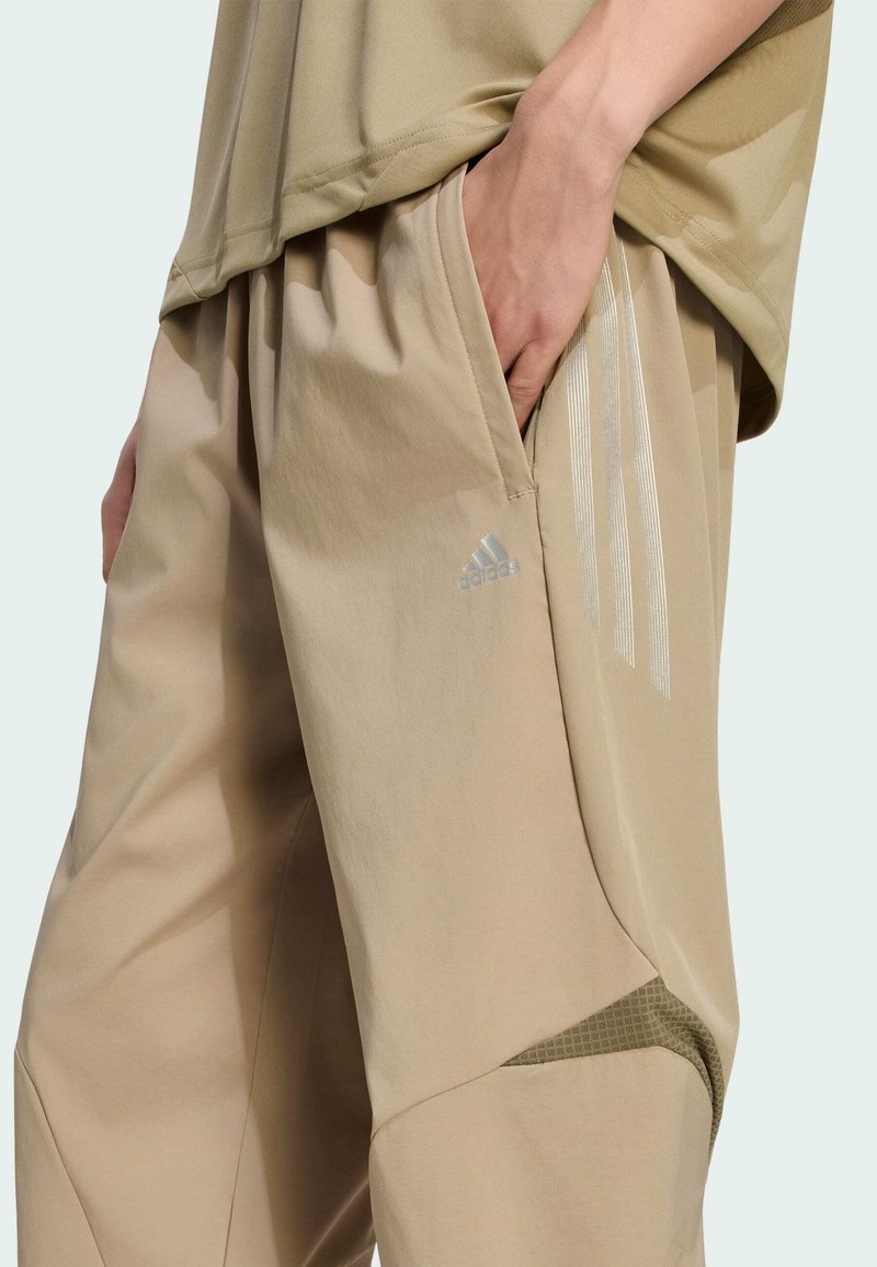 Pantalon de sport beige avec une coupe fuselée, comprenant un accent en mesh, des poches latérales et trois bandes argentées Adidas sur la cuisse.