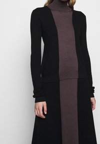 Kvinna iklädd en svart och mörkbrun långärmad polokragsklänning med en vertikal färgblockdesign i mitten.