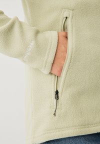 Giacca in fleece verde chiaro con tasca laterale con zip, materiale testurizzato e logo ricamato sulla manica. Visibili il tirante della zip e i dettagli delle cuciture.