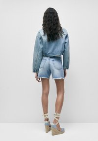 Blå denimcroppad jacka och shorts med fransad fåll, tillsammans med blå espadrillosandaler med beige snören. Enkelt bakgrund.