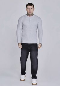 Grå långärmad henley-tröja med uttagen krage, kombinerad med mörka jeans och vita sneakers. Har en figursydd design och synliga tatueringar.