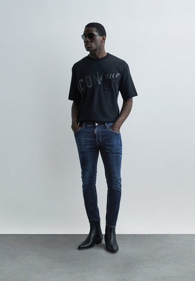 Homme portant des lunettes de soleil noires, un t-shirt noir avec le texte "CONigue", un jean bleu foncé ajusté et des bottines noires à bout pointu, debout les mains dans les poches.