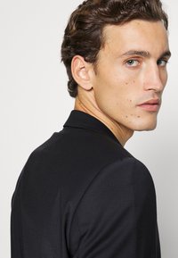 Zwarte, op maat gemaakte_colbert met een gladde textuur, met een notch-lapel en keurig afgewerkte randen. Gezien vanuit een lichte achterhoek.