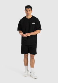 Camiseta de algodón negra de manga corta con un logo en el pecho, combinada con pantalones cortos negros. El modelo lleva zapatillas blancas y calcetines.