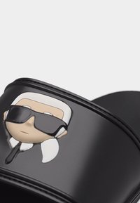 Μαύρο σανδάλι slide με καρτούν πρόσωπο που φοράει λευκά μαλλιά, μαύρα γυαλιά ηλίου και μαύρη γραβάτα στο λουράκι.