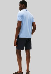 Homme portant un polo bleu clair, un short marine et des mocassins marron, se tenant de dos contre un fond gris uni.