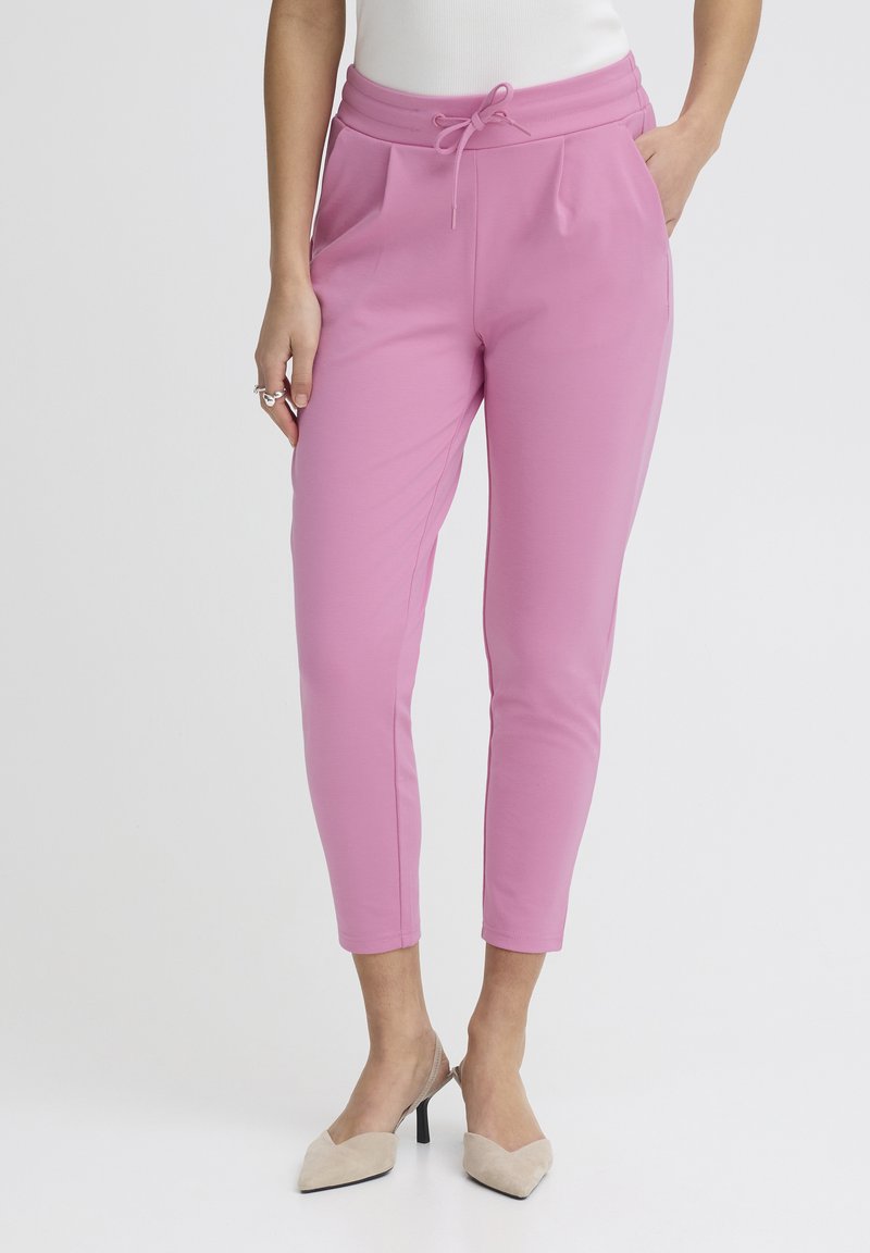 Femme portant un pantalon rose sur mesure, court, avec une taille à cordon, et des escarpins beige à bout pointu et talons kitten, debout avec une main dans la poche.