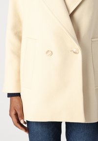 Cappotto beige doppiopetto con una finitura testurizzata, dotato di grandi bottoni chiari e tasche laterali, indossato sopra jeans in denim scuro.