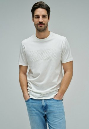 Hombre con cabello oscuro y barba, lleva una camiseta blanca de manga corta con el logo en relieve "Salsa Jeans", jeans azul claro, manos en los bolsillos, fondo neutro.