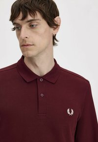 Polo shirt bordeaux in tessuto texture, con due bottoni e un piccolo logo ricamato sul lato sinistro del petto. Presenta un colletto classico.