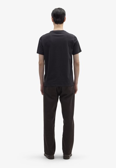 Άντρας που στέκεται με την πλάτη, φορώντας μαύρο κοντομάνικο μπλουζάκι, σκούρο παντελόνι και σκούρα παπούτσια, μπροστά σε απλό λευκό φόντο.