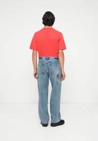 Crvena majica kratkih rukava uparena s širokim plavim trapericama koje imaju zakrčaste džepove i opušteni kroj nogavica, nošena uz crne cipele.