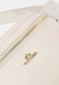 Krémová kožená taška s texturovaným povrchem, vybavená zlatým zipem a zlatým vyraženým logem. Součástí je nastavitelné ramínko.